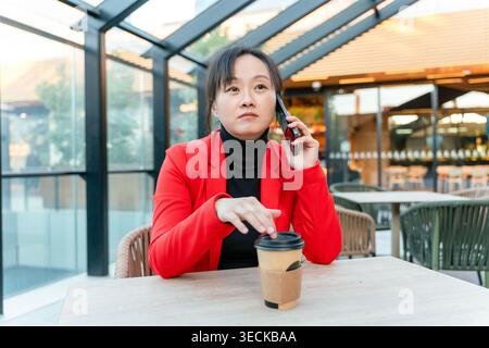 Middle-aged businesswoman in red blazer talks on phone while sitting at a modern cafe with a coffee. The setting is urban with glass walls and outdoor Stock Photo