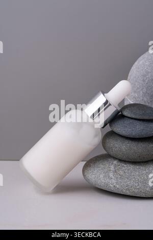 A frosted glass dropper bottle of skincare serum rests elegantly against rounded gray stones. This minimalist setup exudes a serene and clean aestheti Stock Photo