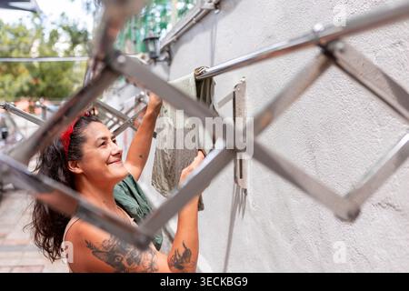 Smiling woman with tattoos hangs clothes on a wall-mounted drying rack outdoors, capturing everyday domestic life, eco-friendly air drying, and home r Stock Photo