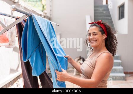 Smiling woman hangs clean laundry on a drying rack in a sunny home patio, enjoying a simple domestic routine, outdoor chores, and everyday sustainable Stock Photo