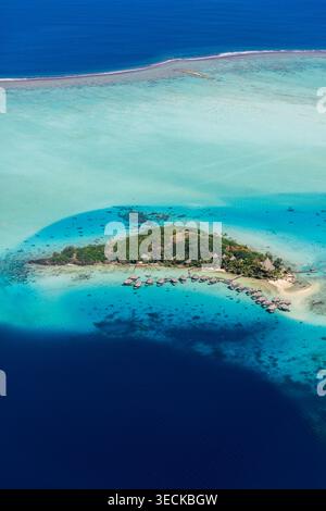 Aerial view of Bora Bora lagoon in French Polynesia, showing turquoise shallows, coral reef line, a small islet with beach huts, and deep blue ocean b Stock Photo