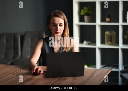 Focused businesswoman working on a laptop at a modern office desk, using a mouse and concentrating on tasks, with shelves and decor in the background. Stock Photo