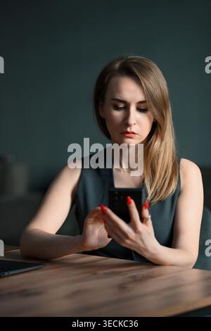 Focused businesswoman working at a desk in a modern office, checking messages on a smartphone beside a laptop, thoughtful and professional in low ligh Stock Photo
