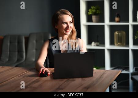 Smiling businesswoman working at a wooden desk with laptop and mouse in a modern office interior, looking aside in natural light, focused on business Stock Photo