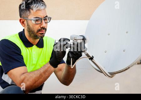 Technician wearing safety vest, gloves and eye protection performs maintenance on a satellite dish connection, ensuring occupational safety during out Stock Photo