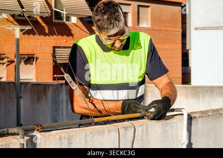 Technician wearing high visibility vest, gloves and safety glasses performs rooftop maintenance, fastening cable and hardware near a building edge in Stock Photo