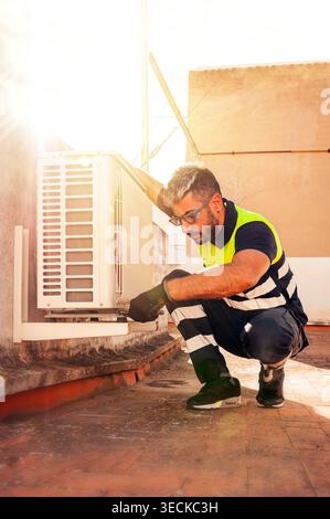 Technician in safety vest and gloves performs outdoor air conditioner maintenance, inspecting and repairing HVAC unit on a rooftop with protective eye Stock Photo
