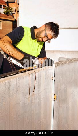 Maintenance technician wearing safety vest, gloves, and protective glasses uses an angle grinder to cut metal on a rooftop wall, demonstrating occupat Stock Photo