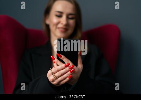 Smiling businesswoman in blazer using a smartphone while sitting in a modern office chair, showcasing mobile communication, remote work, productivity, Stock Photo