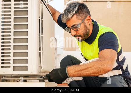 Close-up of a technician in high visibility vest and safety glasses performing maintenance on an outdoor air conditioner unit, using tools and protect Stock Photo