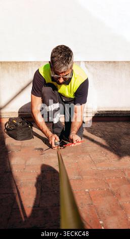 Technician in safety vest kneels on a rooftop terrace, using a measuring tape to measure distance for maintenance, inspection, or construction work in Stock Photo