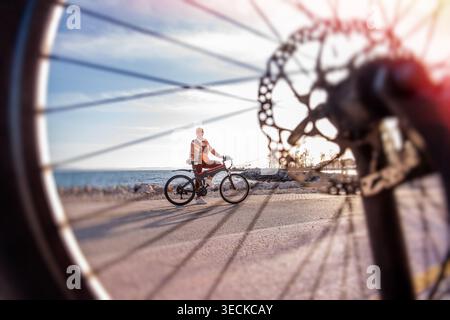 Woman standing with an electric bicycle on a coastal road at sunset, framed through a close-up bike wheel and disc brake, capturing leisure travel and Stock Photo