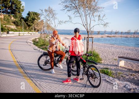 Two sporty women friends take a break on electric bicycles along a seaside path, chatting and checking a phone, enjoying an active outdoor ride on a s Stock Photo