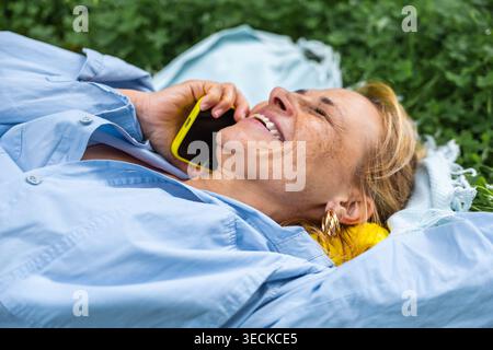 A joyful woman lying on the grass, smiling while speaking on her phone. She enjoys a peaceful outdoor moment, embracing the natural surroundings in le Stock Photo