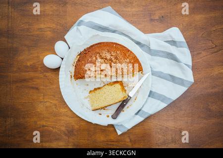 Overhead view of a homemade sponge cake with a slice cut out on a white plate, beside two eggs, a knife, and a striped towel on a rustic wooden table. Stock Photo