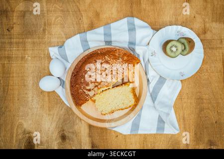 Overhead view of homemade sponge cake with a slice cut out on a wooden board, with eggs, striped kitchen towel, and kiwi slices on a white plate on ru Stock Photo