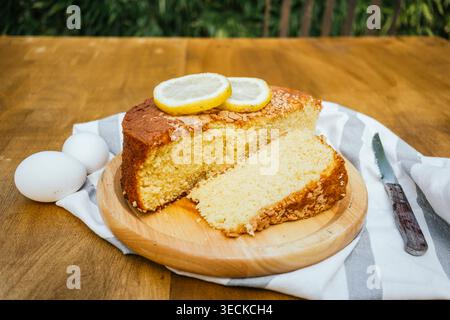 Rustic homemade lemon sponge cake with a sliced piece on a wooden board, topped with lemon rounds, with eggs and a knife on a striped cloth on a table Stock Photo