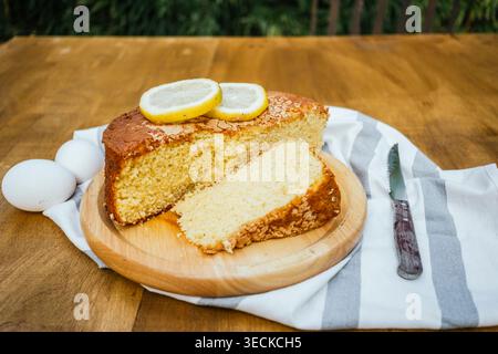 Close-up of homemade lemon sponge cake with a sliced piece on a wooden board, topped with fresh lemon slices, with eggs and a knife on a rustic table. Stock Photo