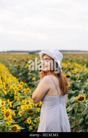 Smiling young woman in a white hat and summer dress stands in a blooming sunflower field at golden hour, enjoying nature and calm countryside atmosphe Stock Photo