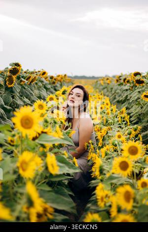 Young woman in a light dress sitting between rows of blooming sunflowers, surrounded by golden petals and green leaves under a soft cloudy sky. Stock Photo