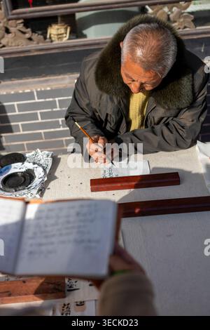 An elderly man practices traditional Chinese calligraphy with brush and ink at an outdoor table, writing characters on paper in winter light, preservi Stock Photo