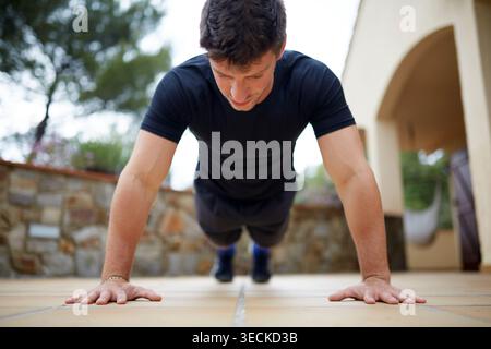 Focused young man doing a push up on a patio during an outdoor workout. Bodyweight exercise for strength, endurance, and healthy lifestyle training in Stock Photo
