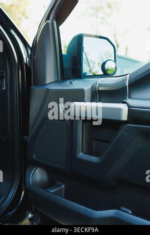 A truck door is open revealing the inside. There are controls on the armrest and a side mirror showing green trees outside in the afternoon light. Stock Photo