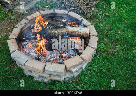 People enjoy warmth from a fire pit in a backyard. The fire burns brightly with flames rising. Grass surrounds the stone structure holding the fire. Stock Photo