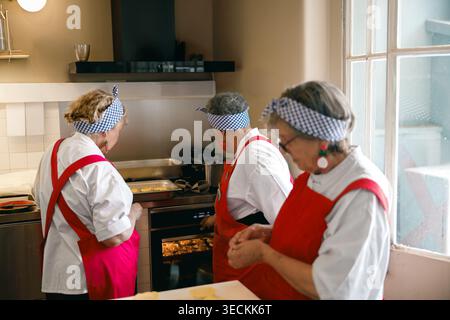 Senior Caucasian women in red aprons baking pastries in a professional kitchen setting Stock Photo