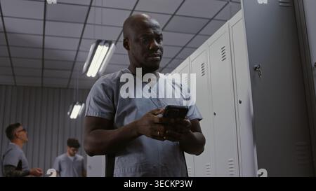 African American Concerned Male Doctor Looking at Phone With Shocked Expression, Appearing Confused and Saddened by Urgent News in Professional Hospital Setting. Concept of Human Side of Medicine. Stock Photo
