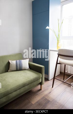Modern living space showcases a green sofa with a decorative pillow, blue cabinetry, and a wooden floor illuminated by natural light from a window Stock Photo