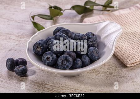 Pile of fresh organic blueberries in elegant white bowl, few berries scattered on light textured wooden surface. Natural light illuminates rich blue p Stock Photo