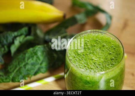 Nutritious vegetable and fruit smoothie in a glass. Focus on the texture of green foam. Banana and kale leaves are blurred in the background on a wood Stock Photo