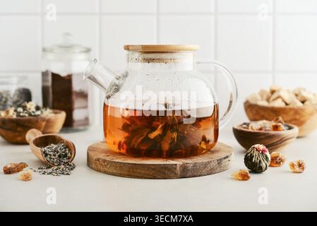 Blooming tea in a glass teapot with dry tea leaves, brown sugar in bowls, and ingredients on a light background. Concept of tea ceremony, natural beve Stock Photo