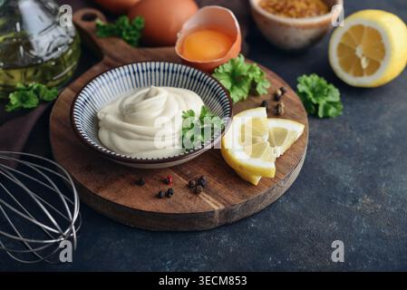 Homemade mayonnaise in a bowl on a wooden board with fresh ingredients including eggs, olive oil, mustard, lemon, parsley, and pepper on a dark backgr Stock Photo
