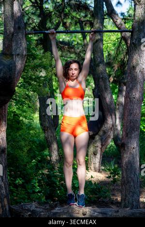 Athletic woman in orange activewear hangs from a pull-up bar suspended between trees, performing a bodyweight exercise outdoors in a sunlit forest par Stock Photo