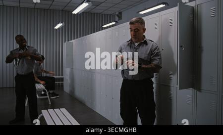 Three Athletic Male Airport Security Officers in Grey Uniforms in Modern Locker Room, Changing Into Civilian Clothes and Chatting, Relaxing After Finishing Work Duty. Concept of Shift Change. Stock Photo