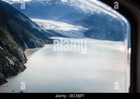 Aerial view through an aircraft window of a massive tidewater glacier flowing into a silty Alaskan fjord surrounded by steep mountains. Stock Photo