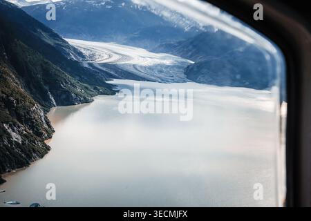 Aerial view through an aircraft window of a massive tidewater glacier flowing into a silty Alaskan fjord surrounded by steep mountains. Stock Photo