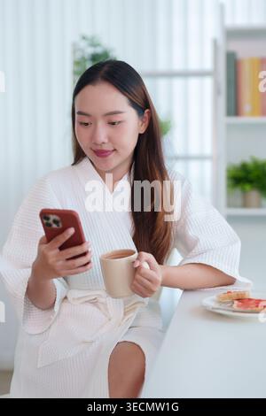Young Asian woman in bathrobe holding coffee cup using smartphone checking social media during morning breakfast Stock Photo