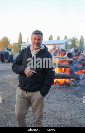 Smiling mature man standing near a large outdoor bonfire during evening festival, enjoying warmth, leisure time and traditional spring celebration. Stock Photo
