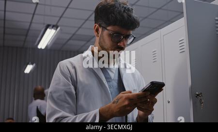 Young Male Physician in White Coat Stands by Locker After Shift, Intensely Reviewing Urgent Laboratory Results and Patient Diagnostics on Smartphone. Concept of Medical Urgency and Dedication. Stock Photo