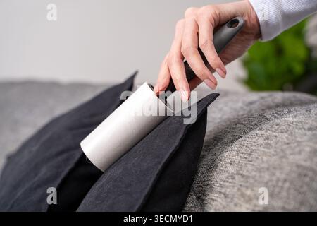 Close-up female hand using sticky lint roller on black fabric cushion over gray sofa removing visible pet hair, dust. Concept of daily soft furniture Stock Photo