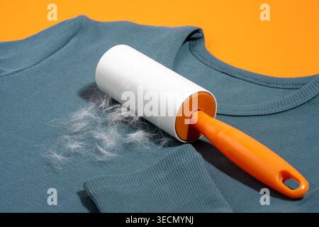 Adhesive lint roller removing pet hair from teal blue sweatshirt on bright orange background. Concept of sticky tape solution for shedding control, fa Stock Photo