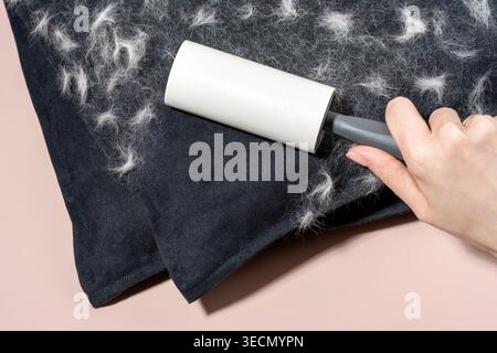 Hand using a lint roller to clean pet hair from a dark fabric surface, showing accumulated lint and fibers on the fabric, set against a light pink bac Stock Photo