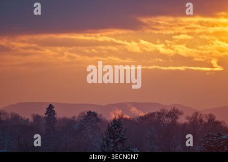 A dramatic low-angle shot of a glowing orange and yellow sunset sky with heavy clouds hanging over a dark, snow-dusted forest and distant mountain pea Stock Photo