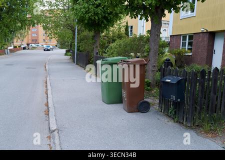 Quiet street lined with trees and residential buildings. Two bins stand beside a fence, one green, one brown. Nynashamn, Stockholm County, Sweden, 23 Stock Photo