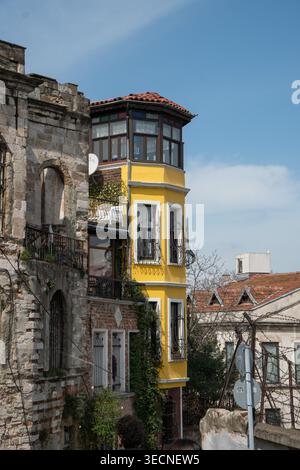 Istanbul, Turkey - April 1, 2026: Yellow corner residential building with bay windows stands next to old stone structure in Balat with tiled roofs Stock Photo