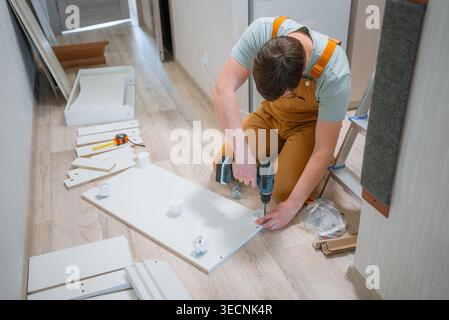 Young man in overalls building white flat-pack furniture at home, using an electric drill to secure parts of a chest of drawers on the floor, surrounded by various assembly components Stock Photo