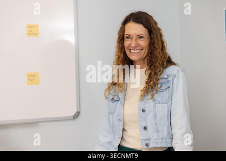 Mature woman in denim standing and smiling, addressing group with two yellow notes on whiteboard Stock Photo